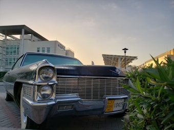 A classic car with an iconic front grille and large headlights is parked on a street. The car has a Dubai license plate and is surrounded by modern architecture, including a building with glass windows and a distinctive overhanging roof. Green foliage is visible in the foreground, and the sky is clear with warm lighting, suggesting a sunset or sunrise.