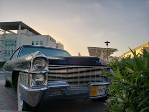 A classic car with an iconic front grille and large headlights is parked on a street. The car has a Dubai license plate and is surrounded by modern architecture, including a building with glass windows and a distinctive overhanging roof. Green foliage is visible in the foreground, and the sky is clear with warm lighting, suggesting a sunset or sunrise.