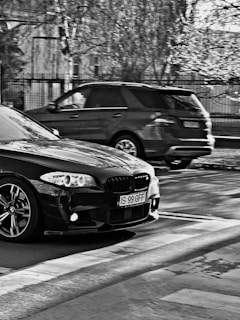 Two cars, one dark-colored sedan in the foreground with a visible license plate, and another SUV in the background, are captured on a city street. The image is in black and white, with trees and a fence visible in the background, hinting at an urban environment.