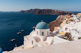 White-washed buildings with blue domes overlook a deep blue sea, with a rocky coastline in the background. Several small boats float on the water near the coast, and the buildings are perched on a hillside, capturing the bright sunlight.