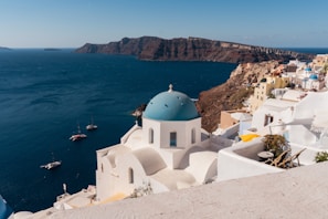 White-washed buildings with blue domes overlook a deep blue sea, with a rocky coastline in the background. Several small boats float on the water near the coast, and the buildings are perched on a hillside, capturing the bright sunlight.