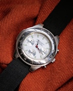 A silver wristwatch with a white dial and multiple sub-dials rests on a textured, orange fabric. The bezel features engraved letters and the watch has a black fabric strap. The watch hands are distinctively styled with a red second hand.
