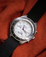 A silver wristwatch with a white dial and multiple sub-dials rests on a textured, orange fabric. The bezel features engraved letters and the watch has a black fabric strap. The watch hands are distinctively styled with a red second hand.