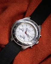 A silver wristwatch with a white dial and multiple sub-dials rests on a textured, orange fabric. The bezel features engraved letters and the watch has a black fabric strap. The watch hands are distinctively styled with a red second hand.