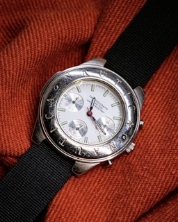 A silver wristwatch with a white dial and multiple sub-dials rests on a textured, orange fabric. The bezel features engraved letters and the watch has a black fabric strap. The watch hands are distinctively styled with a red second hand.