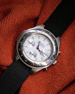 A silver wristwatch with a white dial and multiple sub-dials rests on a textured, orange fabric. The bezel features engraved letters and the watch has a black fabric strap. The watch hands are distinctively styled with a red second hand.