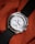 A silver wristwatch with a white dial and multiple sub-dials rests on a textured, orange fabric. The bezel features engraved letters and the watch has a black fabric strap. The watch hands are distinctively styled with a red second hand.