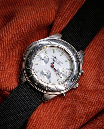 A silver wristwatch with a white dial and multiple sub-dials rests on a textured, orange fabric. The bezel features engraved letters and the watch has a black fabric strap. The watch hands are distinctively styled with a red second hand.