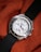 A silver wristwatch with a white dial and multiple sub-dials rests on a textured, orange fabric. The bezel features engraved letters and the watch has a black fabric strap. The watch hands are distinctively styled with a red second hand.