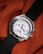 A silver wristwatch with a white dial and multiple sub-dials rests on a textured, orange fabric. The bezel features engraved letters and the watch has a black fabric strap. The watch hands are distinctively styled with a red second hand.