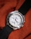 A silver wristwatch with a white dial and multiple sub-dials rests on a textured, orange fabric. The bezel features engraved letters and the watch has a black fabric strap. The watch hands are distinctively styled with a red second hand.