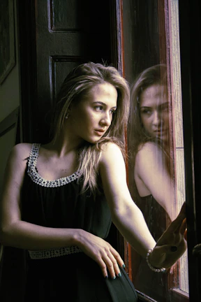 Elegant woman wearing a sleek, modern black dress standing by a sunlit window.