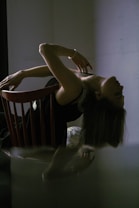 A person leans back on a wooden chair, with their head tilted backwards and one arm gracefully extended. The setting is dimly lit, creating a dramatic shadow effect. The person's attire is dark with light detailing, and they appear to be wearing a bracelet. The overall posture suggests a sense of fluidity and movement, enhanced by soft lighting and reflections.