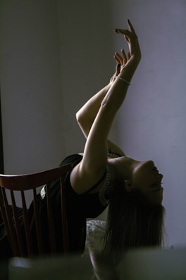 A person leans back on a chair, arching their back with arms gracefully extended upwards. The room is dimly lit, casting a dramatic, shadowy effect.