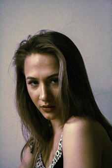 A person with long, dark hair is looking directly at the camera with a serious expression. They are wearing a dark top with white decorative patterns on the straps. The lighting is dim, casting shadows on the face, and the background is plain and muted.