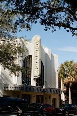 An art deco-style building with the word 'Colony' prominently displayed on its vertical sign. The architecture features tall, narrow windows with decorative elements. Trees with green leaves partially frame the scene, and parked cars are visible in front of the building. The blue sky provides a clear backdrop, enhancing the vintage aesthetic.