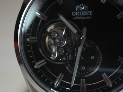 A close-up of an elegant wristwatch featuring a dark dial with silver hour markers and a visible mechanical movement. The brand name 'Orient' is seen near the top, and the hands point towards different times on a small sub-dial.