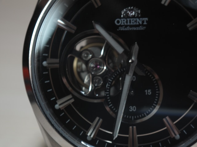 A close-up of an elegant wristwatch featuring a dark dial with silver hour markers and a visible mechanical movement. The brand name 'Orient' is seen near the top, and the hands point towards different times on a small sub-dial.