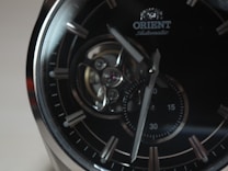 A close-up of an elegant wristwatch featuring a dark dial with silver hour markers and a visible mechanical movement. The brand name 'Orient' is seen near the top, and the hands point towards different times on a small sub-dial.