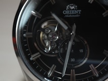 A close-up of an elegant wristwatch featuring a dark dial with silver hour markers and a visible mechanical movement. The brand name 'Orient' is seen near the top, and the hands point towards different times on a small sub-dial.