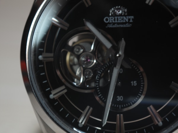 A close-up of an elegant wristwatch featuring a dark dial with silver hour markers and a visible mechanical movement. The brand name 'Orient' is seen near the top, and the hands point towards different times on a small sub-dial.