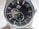A close-up of a luxury wristwatch with a black dial featuring silver hour markers and hands. The watch has a visible mechanism, showcasing its automatic movement. Brand name is visible on the upper part of the dial, with a small sub-dial displaying seconds at the bottom.