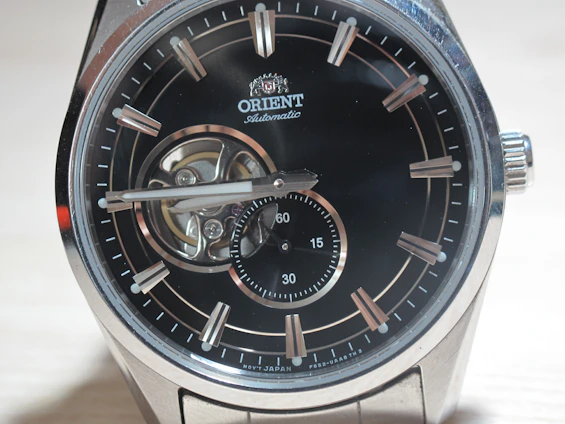 A close-up of a luxury wristwatch with a black dial featuring silver hour markers and hands. The watch has a visible mechanism, showcasing its automatic movement. Brand name is visible on the upper part of the dial, with a small sub-dial displaying seconds at the bottom.