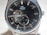 A close-up of a luxury wristwatch with a black dial featuring silver hour markers and hands. The watch has a visible mechanism, showcasing its automatic movement. Brand name is visible on the upper part of the dial, with a small sub-dial displaying seconds at the bottom.