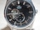 A close-up of a luxury wristwatch with a black dial featuring silver hour markers and hands. The watch has a visible mechanism, showcasing its automatic movement. Brand name is visible on the upper part of the dial, with a small sub-dial displaying seconds at the bottom.