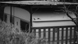 A black and white photograph features a vintage caravan partially obscured by tall grass and a metal fence. The caravan has a sleek, curved roof with a small animal silhouette art on the side. There’s a canopy overhead and leafless branches extending into the frame, giving a secluded and quiet atmosphere.
