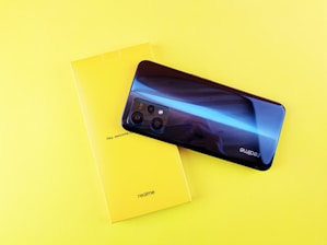 A smartphone with a shiny blue and purple back is placed on top of a bright yellow box displaying the brand 'realme' along with some text. The phone has a reflective surface, showing a partial reflection, and features multiple camera lenses.