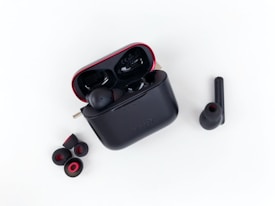 A pair of wireless earbuds with a charging case is displayed. The charging case is black with red accents and is open, showing one earbud inside. Additional ear tips are placed next to the case on a white background.