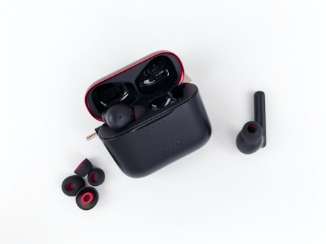 A pair of wireless earbuds with a charging case is displayed. The charging case is black with red accents and is open, showing one earbud inside. Additional ear tips are placed next to the case on a white background.