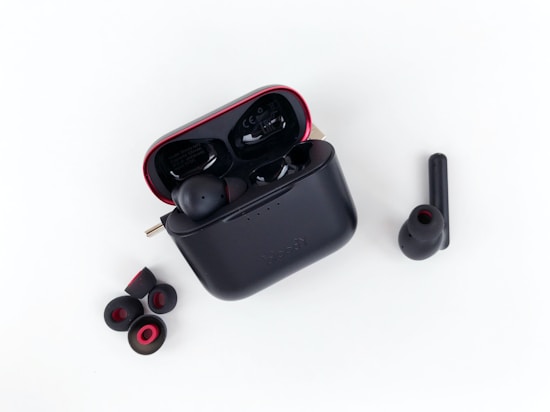 A pair of wireless earbuds with a charging case is displayed. The charging case is black with red accents and is open, showing one earbud inside. Additional ear tips are placed next to the case on a white background.