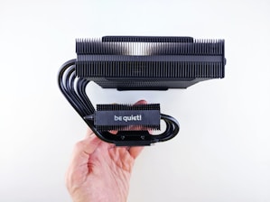 A hand holding a black CPU cooler with multiple metal heat sink fins and several curved heat pipes. The brand name 'be quiet!' is visible on the side of the cooler.