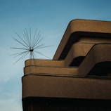 A modern building features smooth, curved lines with a minimalistic aesthetic. The structure is made of concrete with a textured surface. Protruding from the top is a unique antenna or sculpture composed of multiple metal rods radiating outward, set against a clear blue sky.