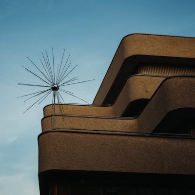A modern building features smooth, curved lines with a minimalistic aesthetic. The structure is made of concrete with a textured surface. Protruding from the top is a unique antenna or sculpture composed of multiple metal rods radiating outward, set against a clear blue sky.