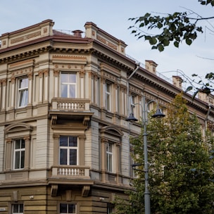 A historic multi-story building with ornate architectural details, including decorative columns and moldings. It features large windows and a small balcony on the corner. The building is surrounded by trees, and there is a street lamp nearby. The sky is clear, creating a serene urban atmosphere.