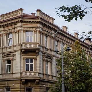 A historic multi-story building with ornate architectural details, including decorative columns and moldings. It features large windows and a small balcony on the corner. The building is surrounded by trees, and there is a street lamp nearby. The sky is clear, creating a serene urban atmosphere.