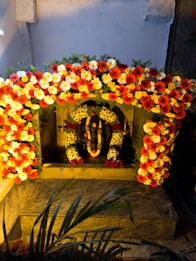 A shrine adorned with vibrant garlands of orange, yellow, and white flowers surrounds a statue of a deity. The scene is set within a stone alcove, with greenery adding a natural touch above the stone structure. The statue is decorated with additional floral garlands, enhancing the religious and festive atmosphere.
