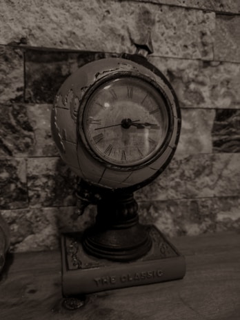A vintage globe clock with Roman numerals placed against a stone wall. The clock has a distressed, antique finish and is mounted on a base labeled 'The Classic.' The background features a textured pattern of differently sized stone tiles, adding to the aged aesthetic.