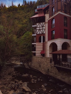 A multi-story building with a red brick facade and timber framing stands by a narrow, rocky river. Tall evergreen trees cover the surrounding hills, contributing to a serene and secluded atmosphere.