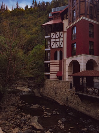A multi-story building with a red brick facade and timber framing stands by a narrow, rocky river. Tall evergreen trees cover the surrounding hills, contributing to a serene and secluded atmosphere.