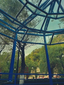 A geometric metal and glass structure forms an overhead canopy, with blue metal beams intersecting at various angles. In the background, lush green trees and well-maintained shrubs create a natural setting. Several globe-shaped street lamps are visible, interspersed among the trees, and a hint of mountainous landscape is present in the distance.