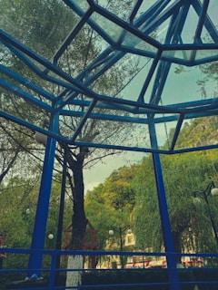 A geometric metal and glass structure forms an overhead canopy, with blue metal beams intersecting at various angles. In the background, lush green trees and well-maintained shrubs create a natural setting. Several globe-shaped street lamps are visible, interspersed among the trees, and a hint of mountainous landscape is present in the distance.
