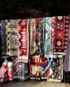 A collection of colorful, intricately patterned rugs with geometric designs hangs vertically. The rugs display a variety of colors including red, brown, black, and blue, and appear to be handmade with traditional motifs. Positioned towards the left, a few plush toys are visible, adding a playful element to the scene.