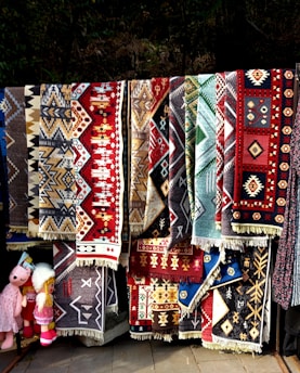 A collection of colorful, intricately patterned rugs with geometric designs hangs vertically. The rugs display a variety of colors including red, brown, black, and blue, and appear to be handmade with traditional motifs. Positioned towards the left, a few plush toys are visible, adding a playful element to the scene.