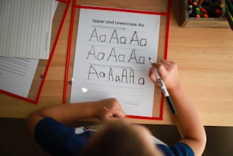 A child is practicing writing the uppercase and lowercase letter 'A' on a worksheet using a black marker. The sheet includes guidelines for tracing the letter. Other worksheets and a box of colored pencils are visible on the wooden table, suggesting an educational environment.