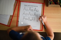 A child is practicing writing the uppercase and lowercase letter 'A' on a worksheet using a black marker. The sheet includes guidelines for tracing the letter. Other worksheets and a box of colored pencils are visible on the wooden table, suggesting an educational environment.