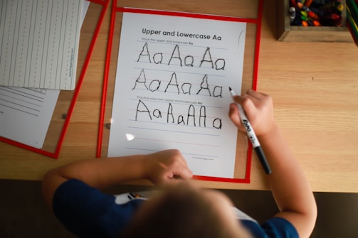 A child is practicing writing the uppercase and lowercase letter 'A' on a worksheet using a black marker. The sheet includes guidelines for tracing the letter. Other worksheets and a box of colored pencils are visible on the wooden table, suggesting an educational environment.